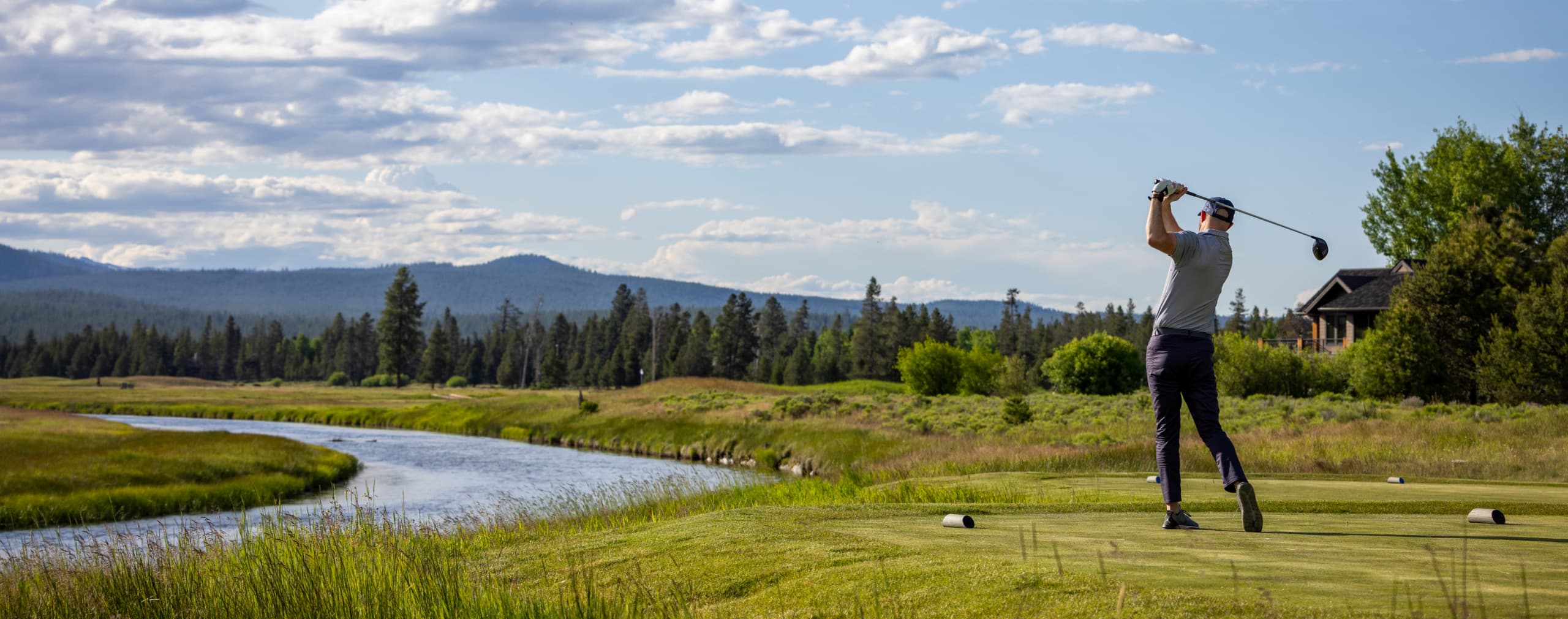 A golfer swings a club on a lush course beside a winding river, with trees, grass, and distant mountains under a partly cloudy sky.
