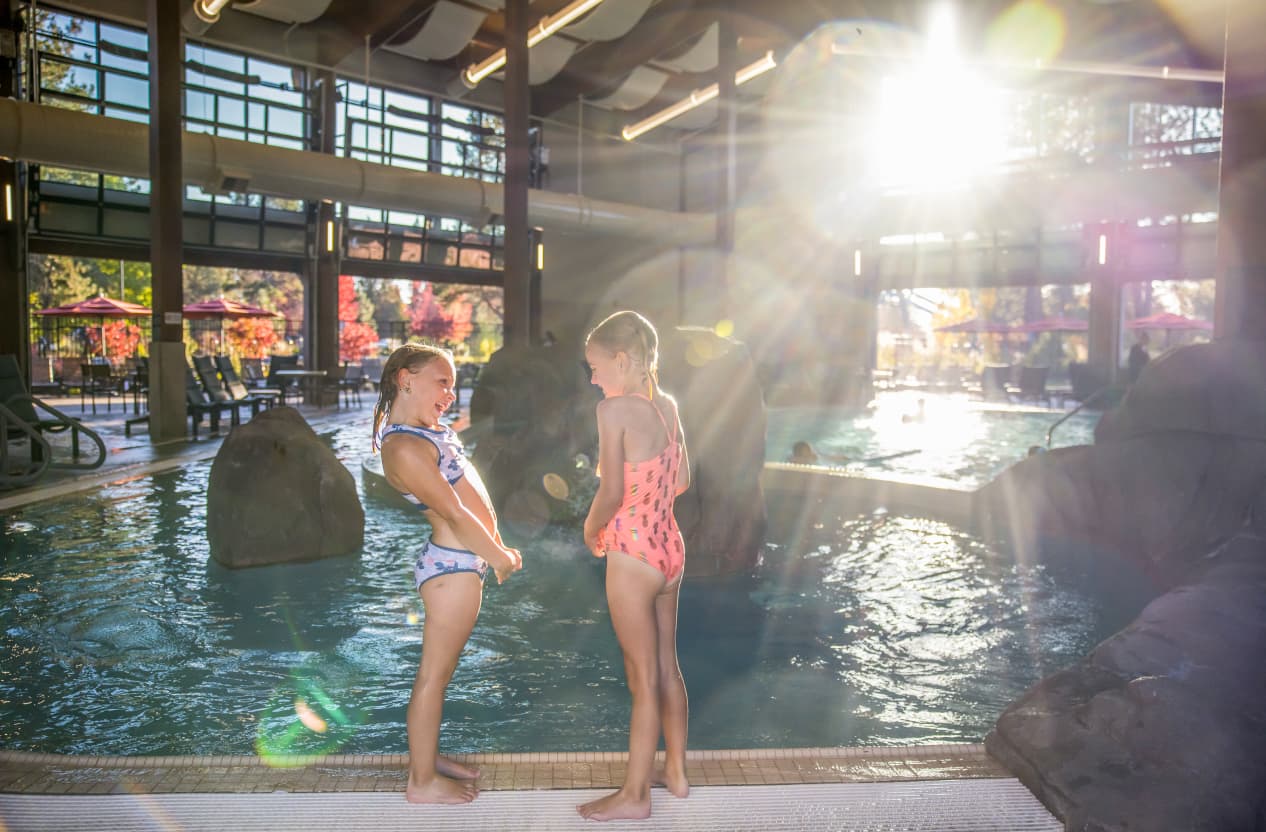 Two children in swimsuits stand at the edge of an indoor pool, smiling at each other as sunlight streams in through large windows, creating a bright, cheerful atmosphere.