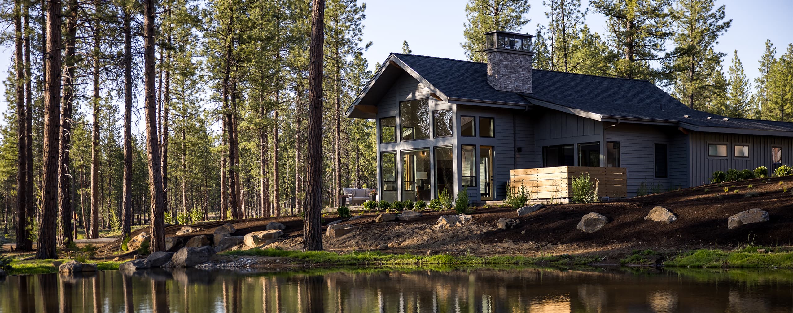A modern cabin with large windows sits beside a small pond, surrounded by tall pine trees and lush greenery on a sunny day.