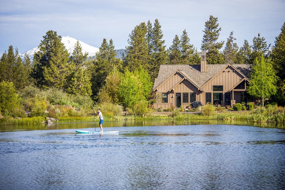 A person paddleboarding on a calm lake in front of a house surrounded by trees, with a snow-capped mountain visible in the background.