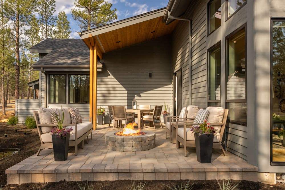 Modern patio with cushioned seating around a stone fire pit, two planters with flowers, and a dining table under a wooden roof, surrounded by trees and a gray house exterior.