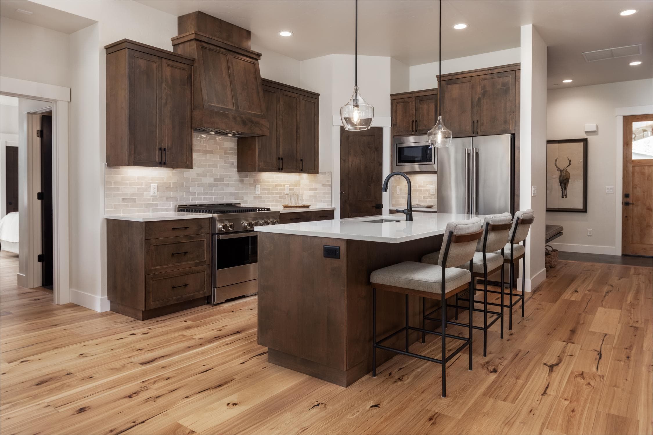 A Forestbrook Home with a modern kitchen with wooden cabinets, stainless steel appliances, an island with three cushioned stools, two pendant lights, light wood floors, and a neutral color palette.