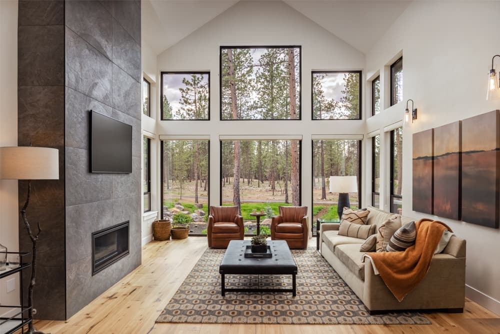 Modern living room with large windows, forest view, neutral-toned furniture, stone fireplace, wall-mounted TV, patterned rug, and orange throw blanket on the sofa. Two leather chairs face the coffee table.