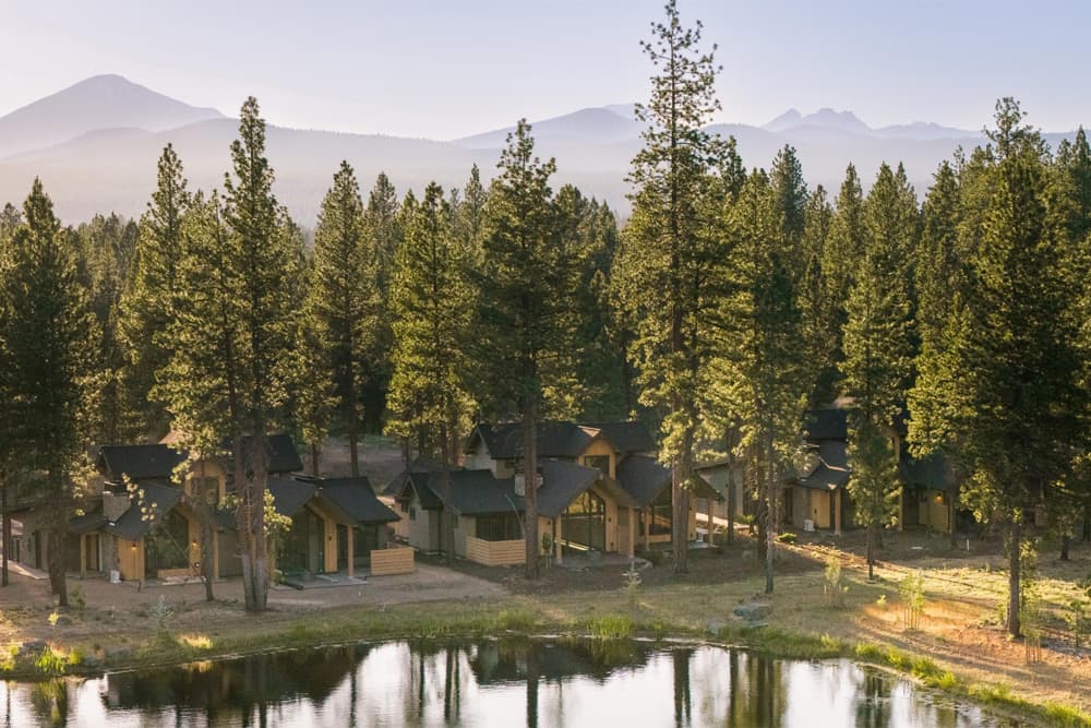 A cluster of Lakeside Homes nestled among tall pine trees beside a tranquil pond, with mountain peaks visible in the background under a clear sky in Caldera Springs overlooking Mt Bachelor.