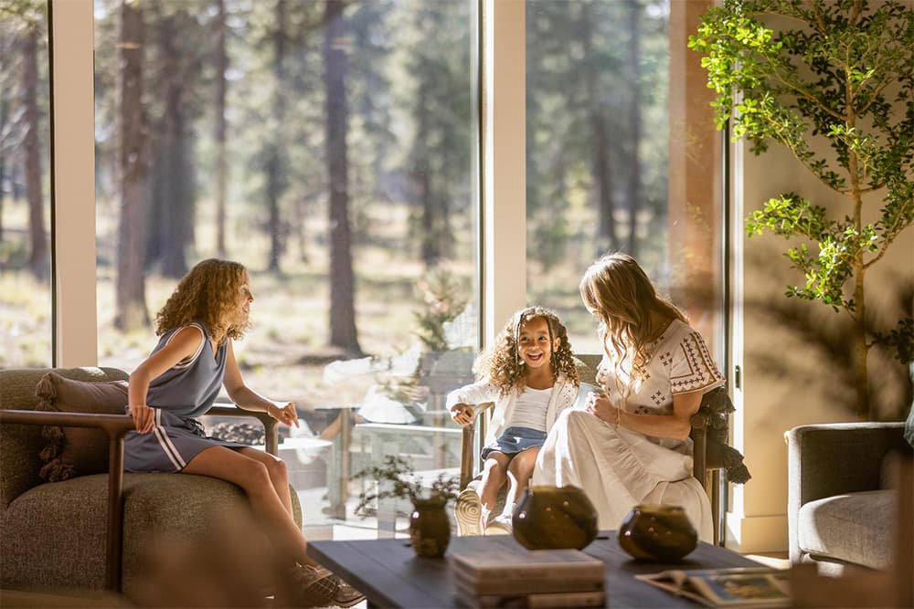 Lakeside at Caldera Springs. Three people sit in a sunlit living room with large windows, smiling and chatting. Tall trees and greenery can be seen outside. The scene feels warm, relaxed, and filled with natural light.