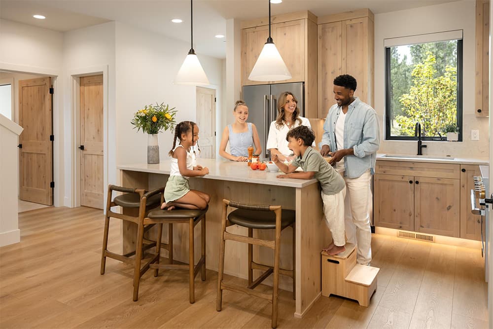 Lakeside at Caldera Springs. A family of five smiles and chats together around a kitchen island in a modern, wood-accented kitchen. Two children sit and stand on chairs while the adults and an older child stand nearby.