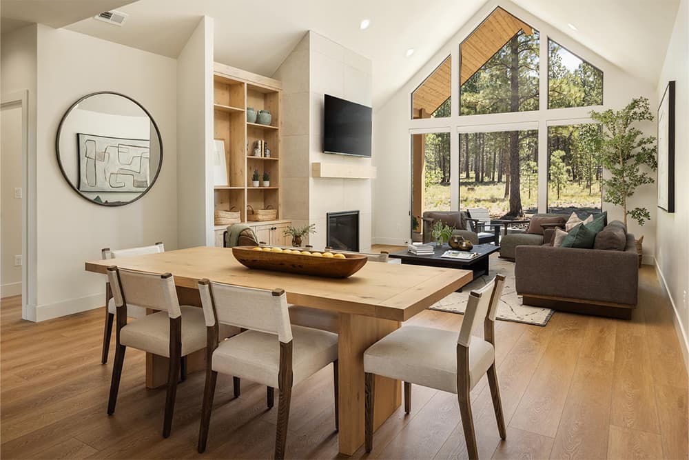 Lakeside at Caldera Springs. Modern open living and dining room with a wooden table, six chairs, beige sofa, large windows showing trees, a TV above a fireplace, shelves, and indoor plants.