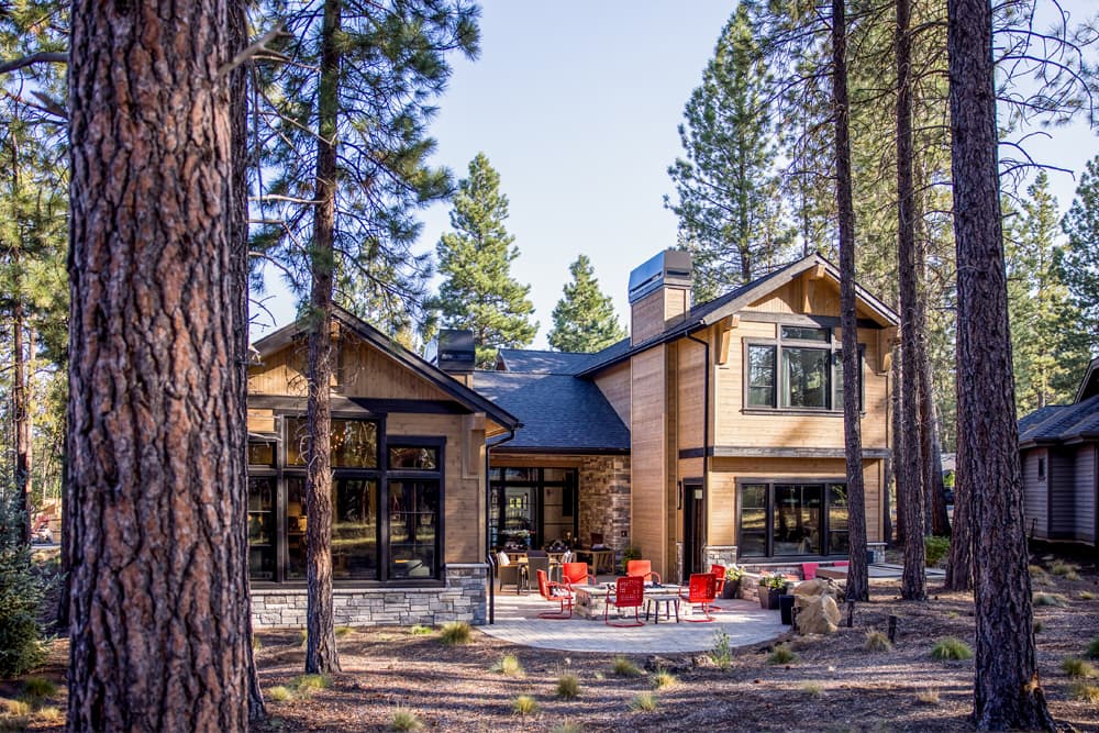 A modern wood cabin with large windows sits among tall pine trees. A patio with red chairs and a fire pit is in the backyard. Sunlight filters through the forest.
