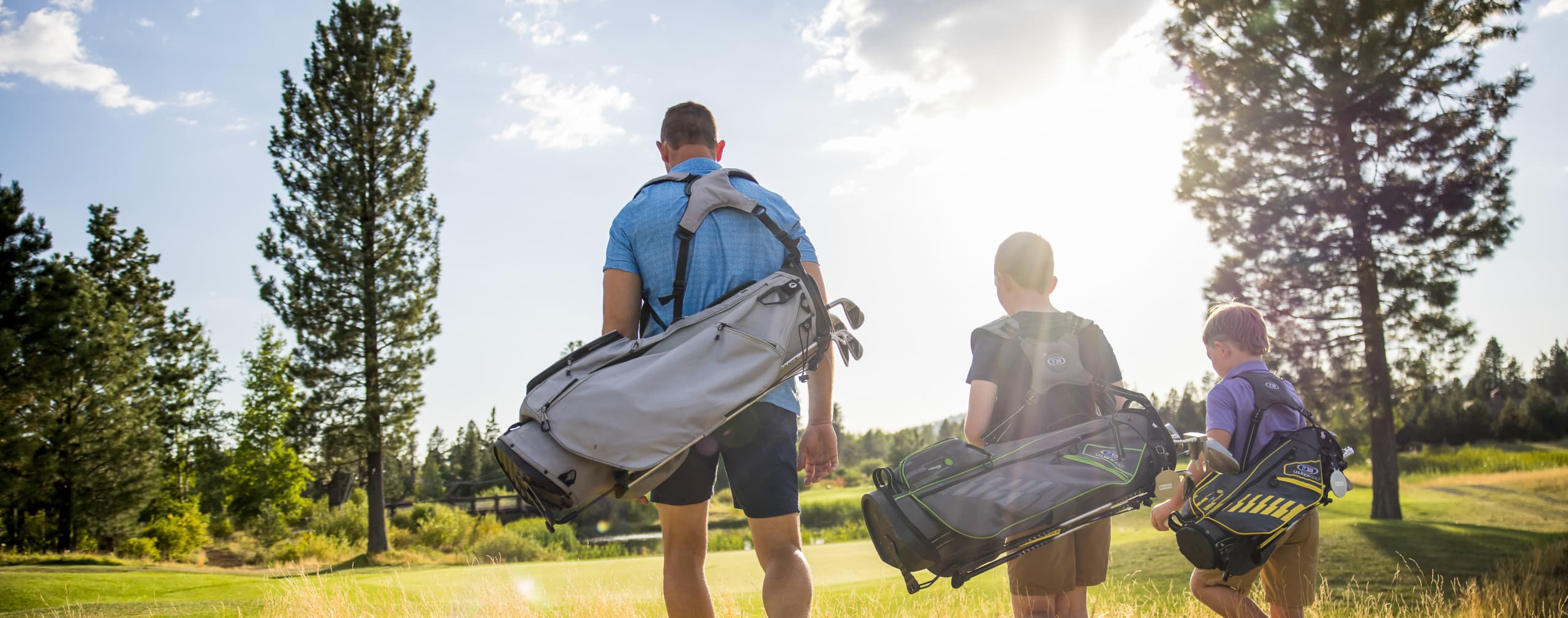 A man and two children walk on a sunny golf course carrying golf bags, surrounded by trees and green grass.