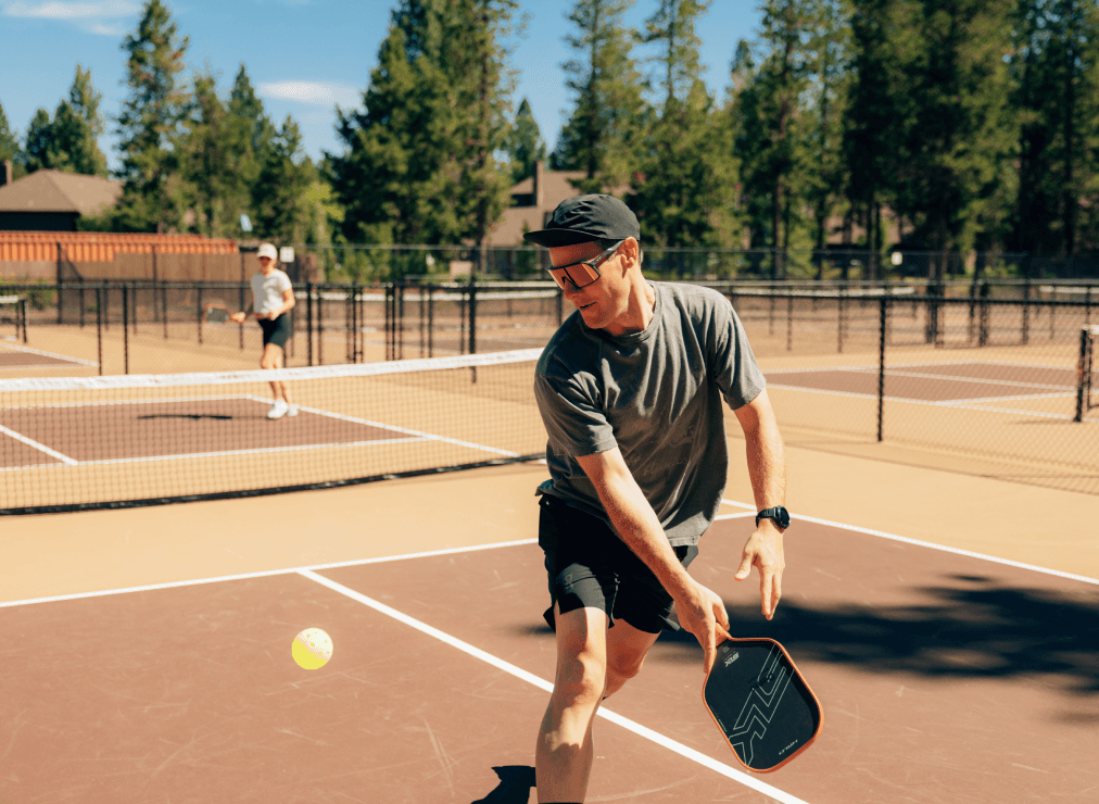 Two people play pickleball on an outdoor court. One player in the foreground prepares to hit the ball with a paddle, while the other stands near the net. Trees and buildings are visible in the background under a sunny sky.