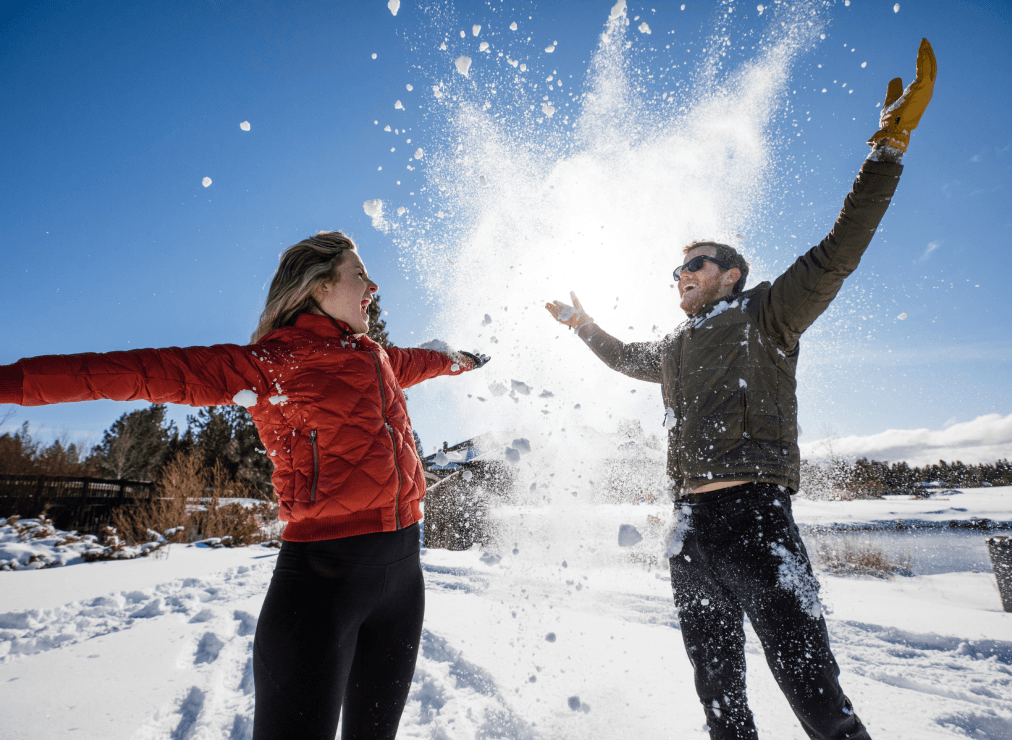 Two people in winter jackets and gloves throw snow into the air, smiling with arms raised, on a sunny, snowy landscape with trees and blue sky in the background.