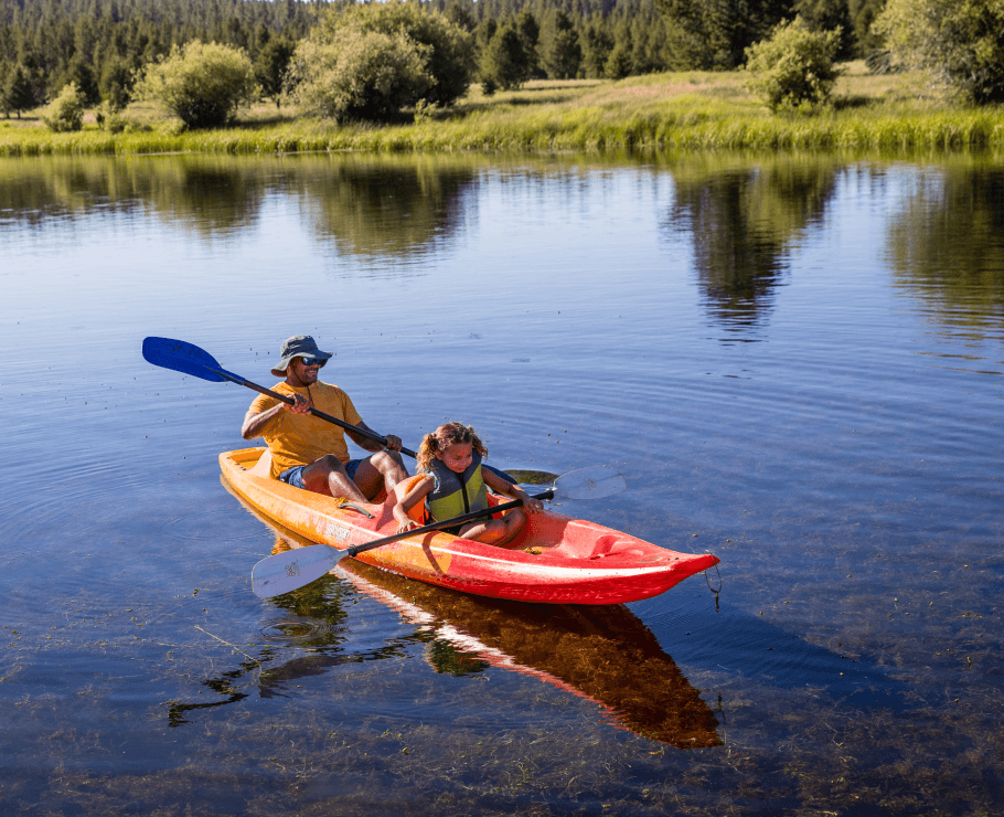 An adult and child paddle a bright orange kayak together on a calm lake, surrounded by green grass and trees under a clear sky.