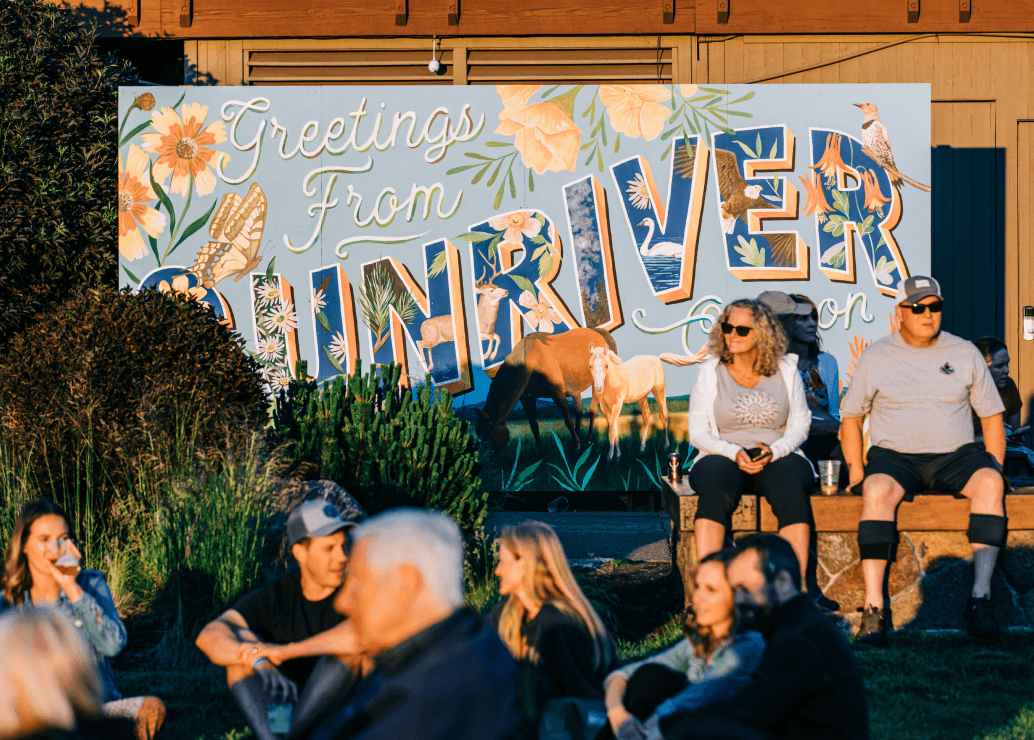 People sit and relax in front of a large colorful sign reading Greetings From Sunriver decorated with flowers, animals, and leaves on a sunny day.