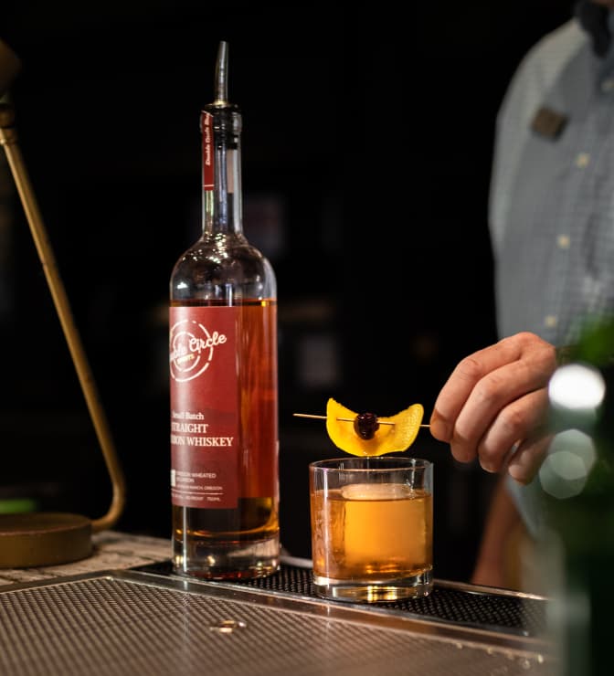 A hand places a garnish of peach and a cherry on a whiskey cocktail beside a tall bottle of whiskey labeled Circle. The scene is set on a dimly lit bar counter.