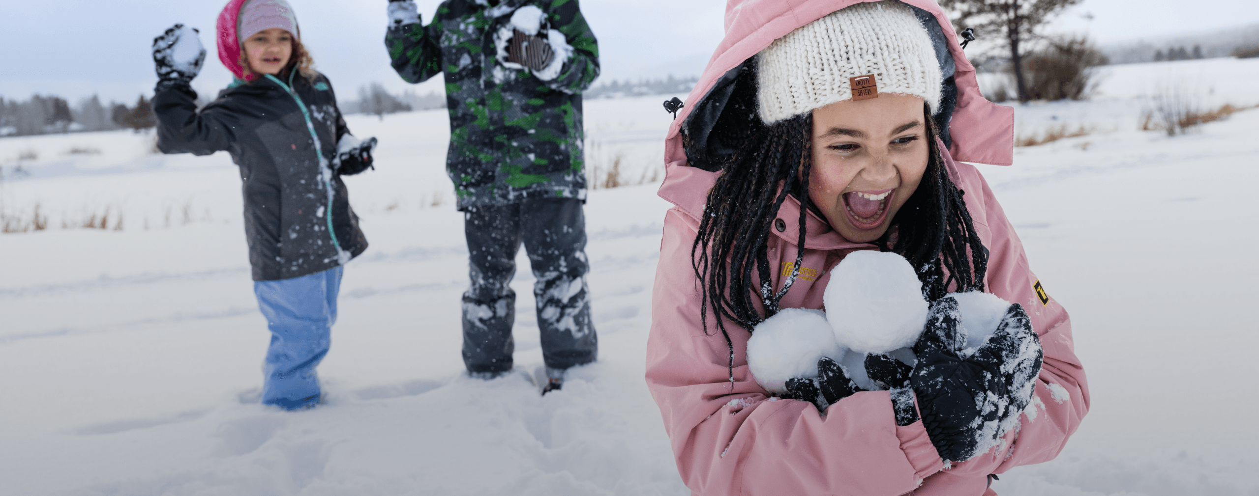 Three children play in the snow; one girl in a pink jacket holds snowballs and laughs, while two other children in winter clothes stand behind her, also enjoying the snowy outdoor scene.