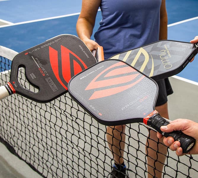 Three people standing on a pickleball court tap their paddles together over the net, possibly before or after a game. One person wears a purple shirt and holds a paddle with a gold design.
