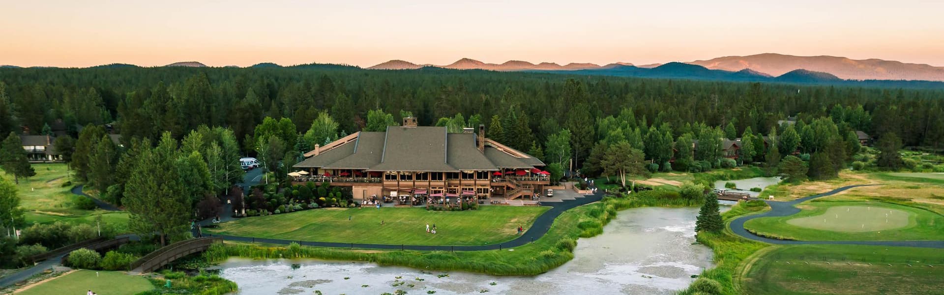 Large lodge-style building surrounded by trees, with a pond and golf course in the foreground and mountains in the background at sunset.