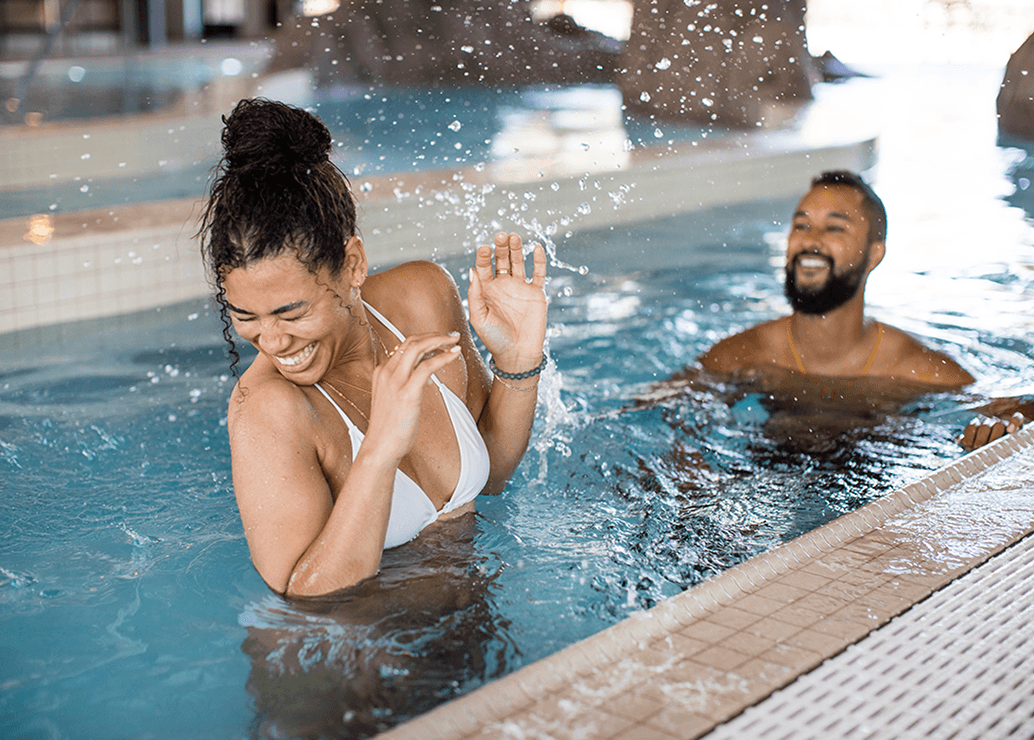 A woman in a white bikini laughs and shields herself from splashing water in a pool, while a man nearby smiles joyfully. The scene is bright and playful, suggesting they are having fun together.