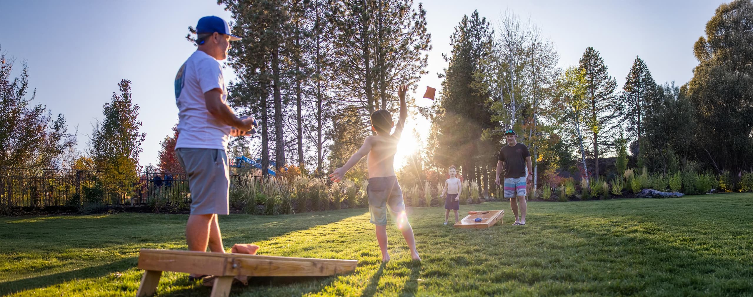 Four people play cornhole on a grassy lawn at sunset, surrounded by tall trees and greenery. One person is tossing a bean bag while others watch, enjoying the outdoor activity.
