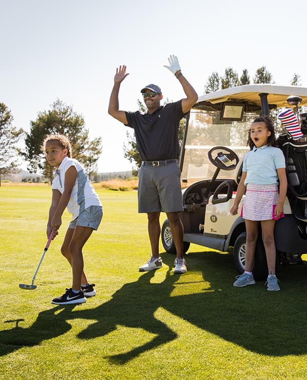 A man stands by a golf cart with his arms raised while two young girls play golf beside him on a sunny day. The girl in front is putting, and the other girl looks surprised.