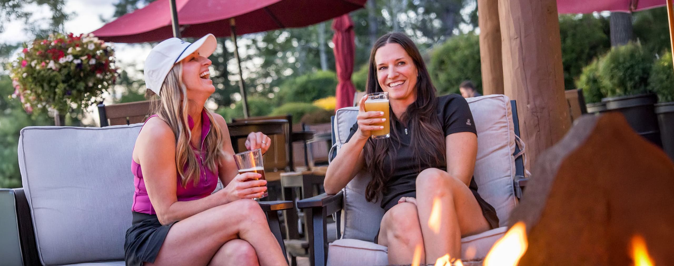 Two women sitting outdoors by a fire pit, smiling and holding drinks. Patio furniture, red umbrellas, and greenery are visible in the background on a sunny day.