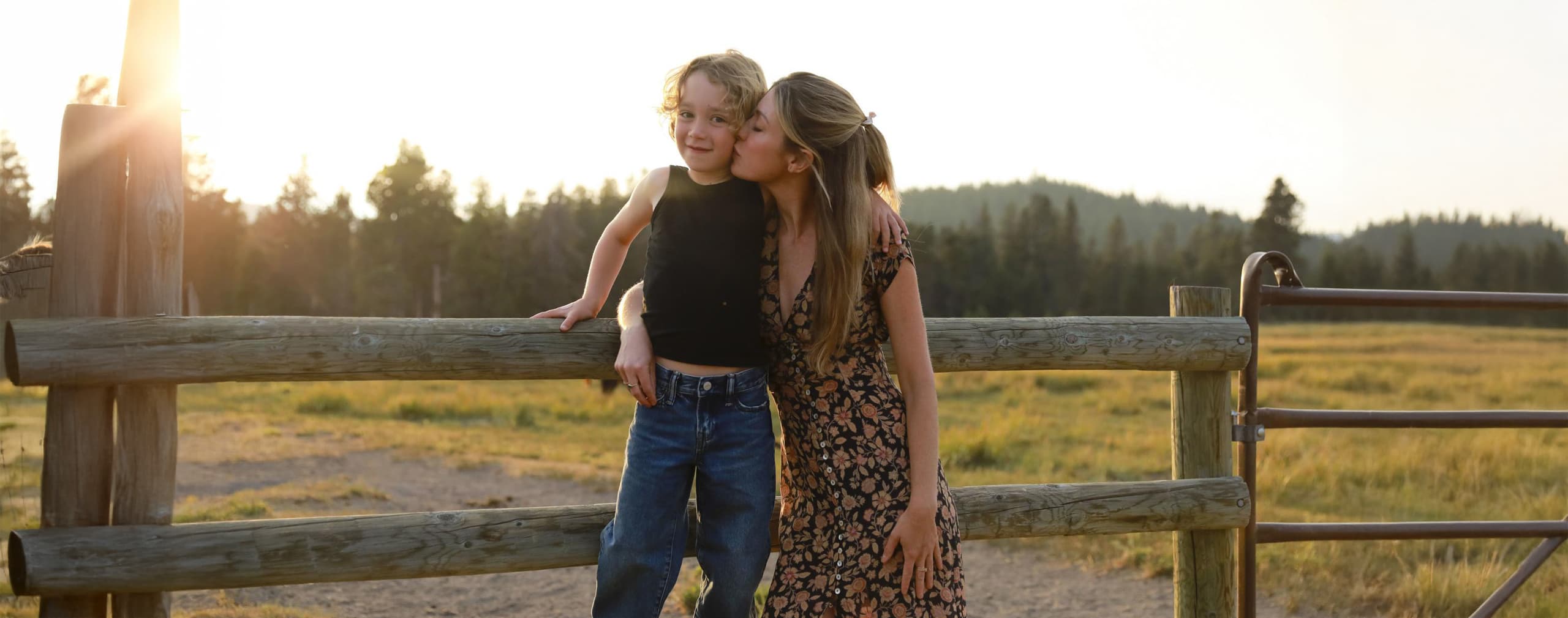 A woman in a floral dress kisses a smiling child on the cheek as they sit together on a wooden fence in a sunlit field with trees in the background.