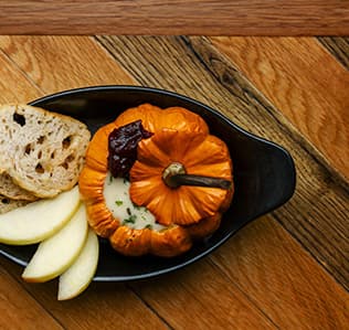 A small baked pumpkin filled with cheese and herbs, topped with a dollop of cranberry sauce, is served in a black dish with slices of apple and pieces of rustic bread on a wooden table.