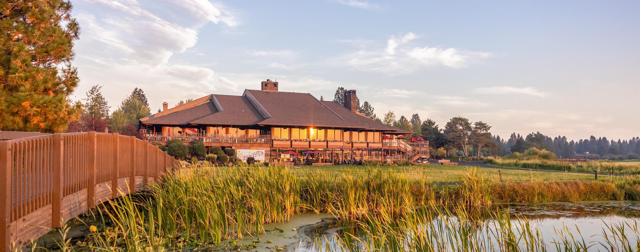 A large lodge-style building with a deck overlooks a pond and green lawn, surrounded by trees under a blue sky at sunset.