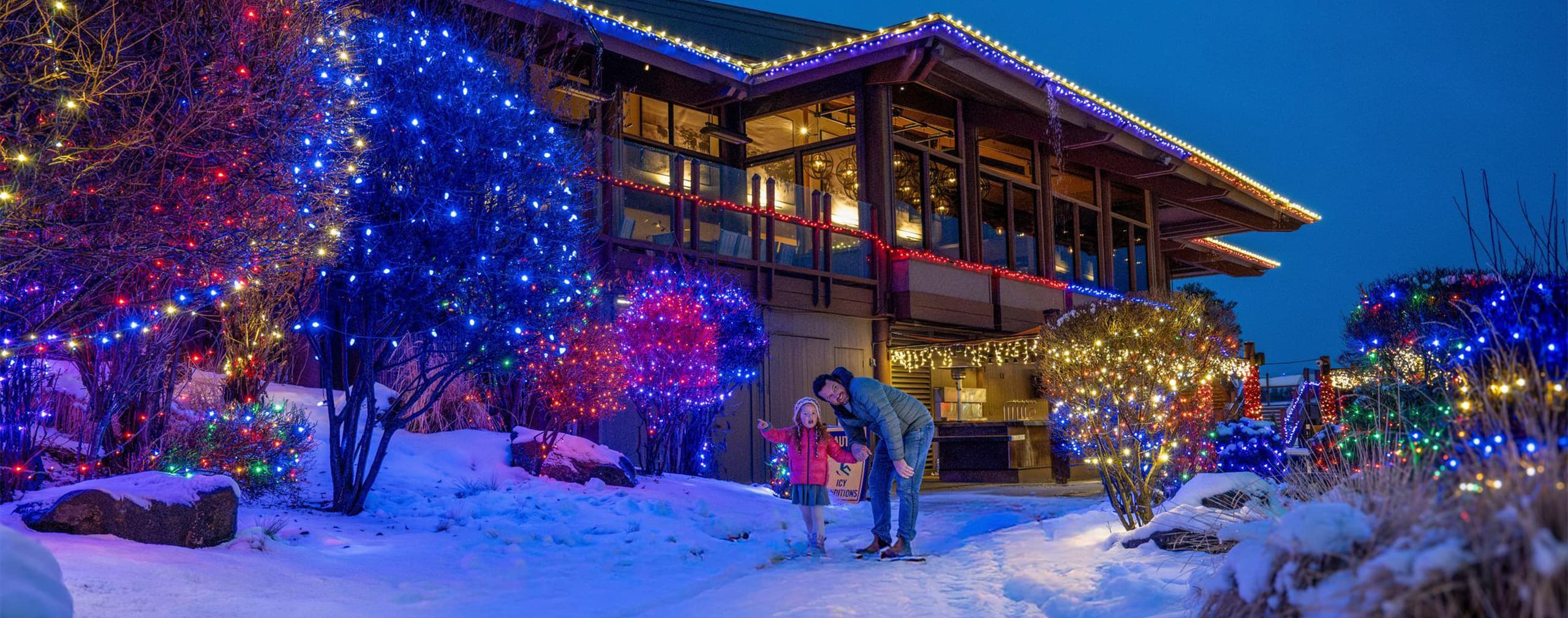A father and child stand on a snowy path outside a building decorated with colorful holiday lights at dusk. The trees and bushes are also covered in bright, festive lights.