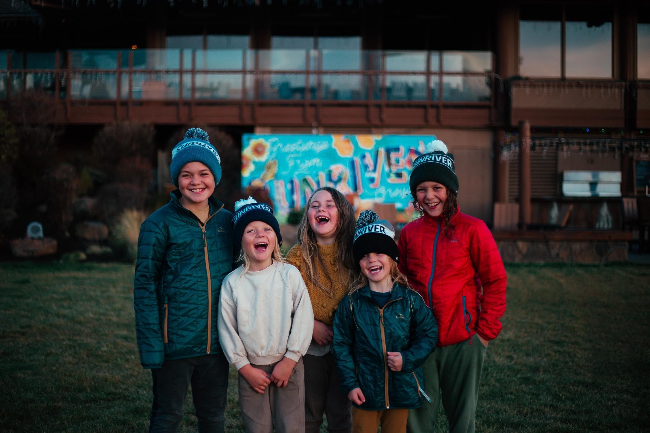 Five children wearing SURVIVOR beanies stand smiling and laughing on grass, with a colorful SURVIVOR sign and a building in the background.