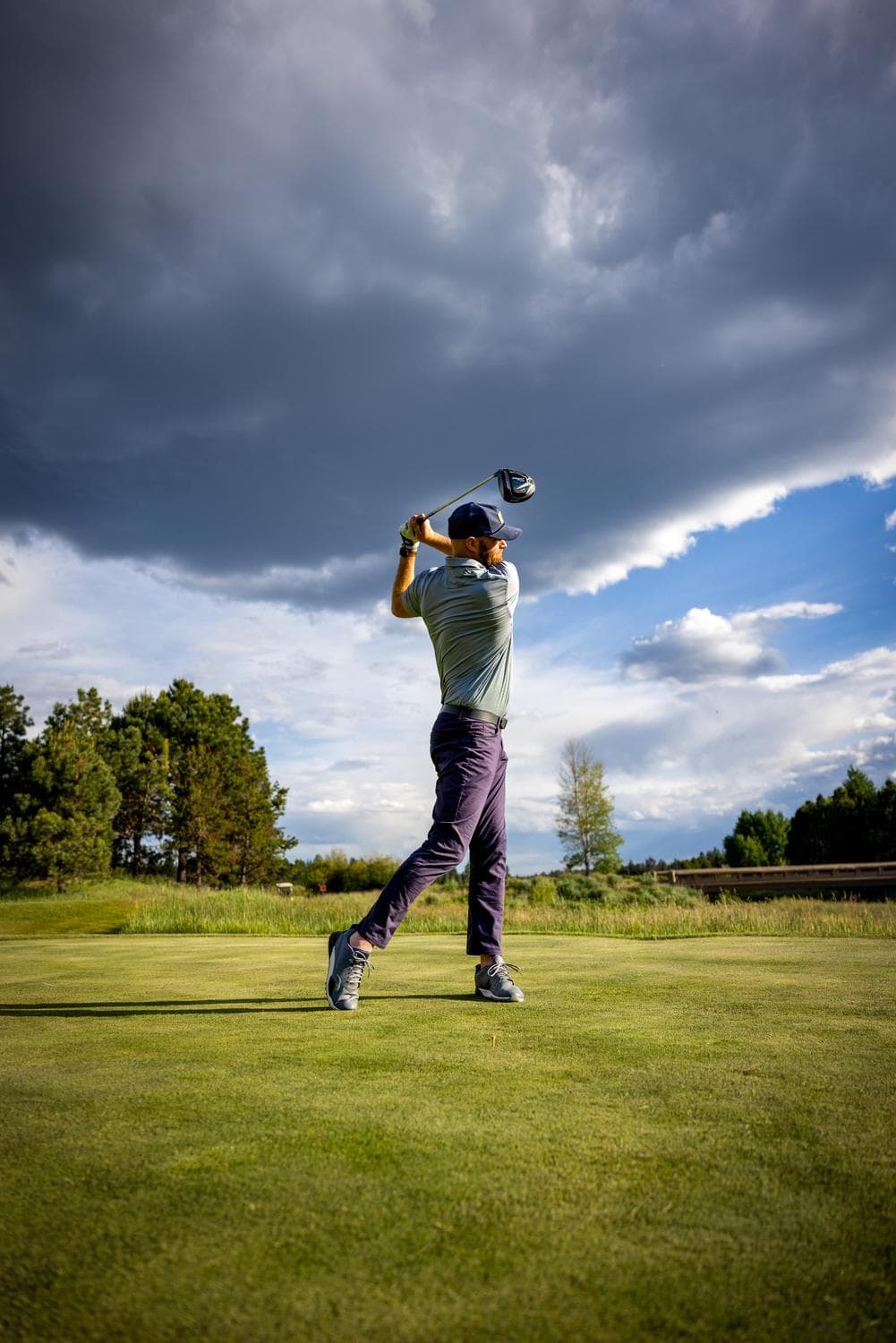A golfer swings a club on a green golf course under a dramatic sky with dark clouds and patches of blue. Trees and grassy landscape are visible in the background.