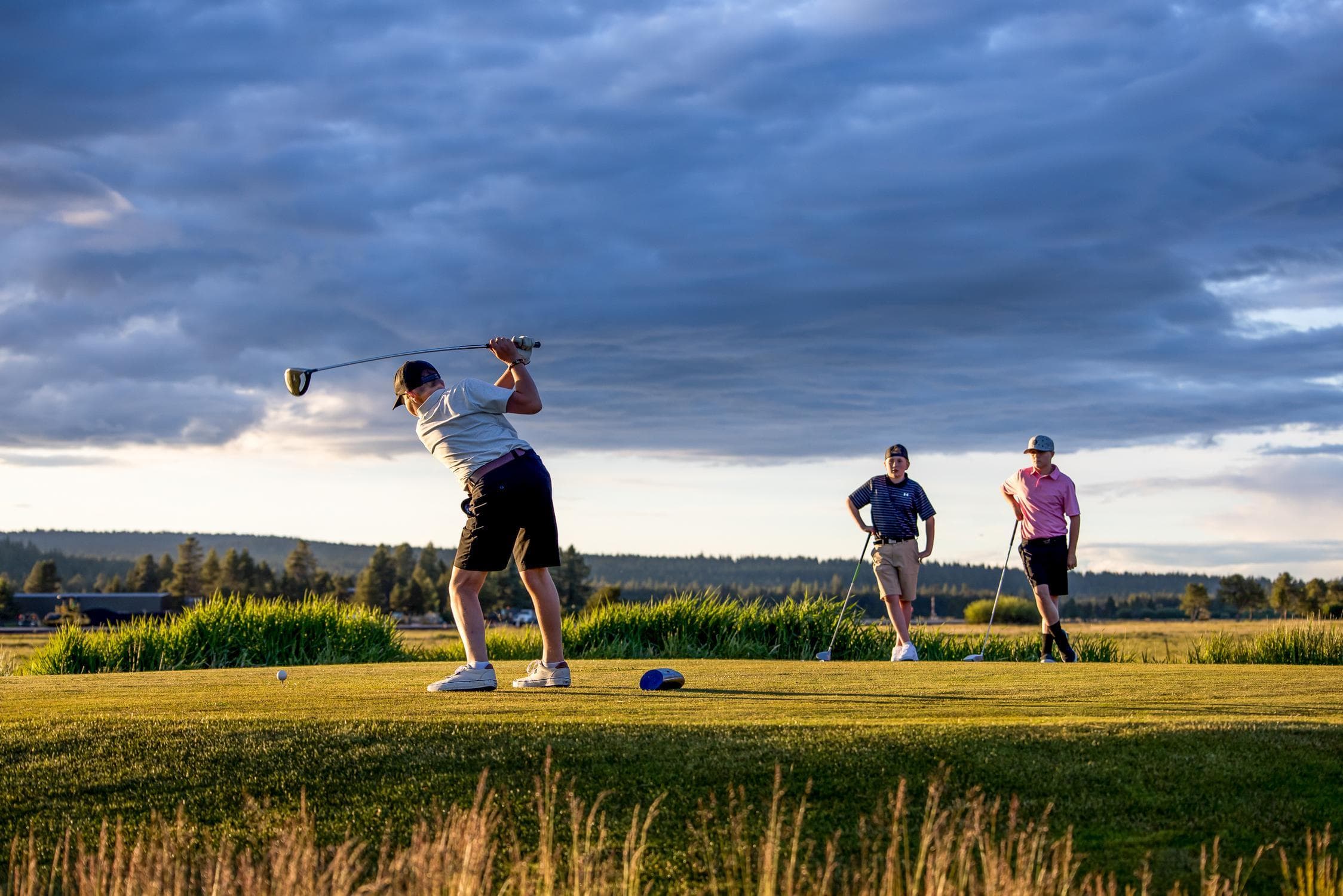 Three people on a golf course at sunset; one is swinging a club while the other two watch. The sky is partly cloudy with trees in the distance.
