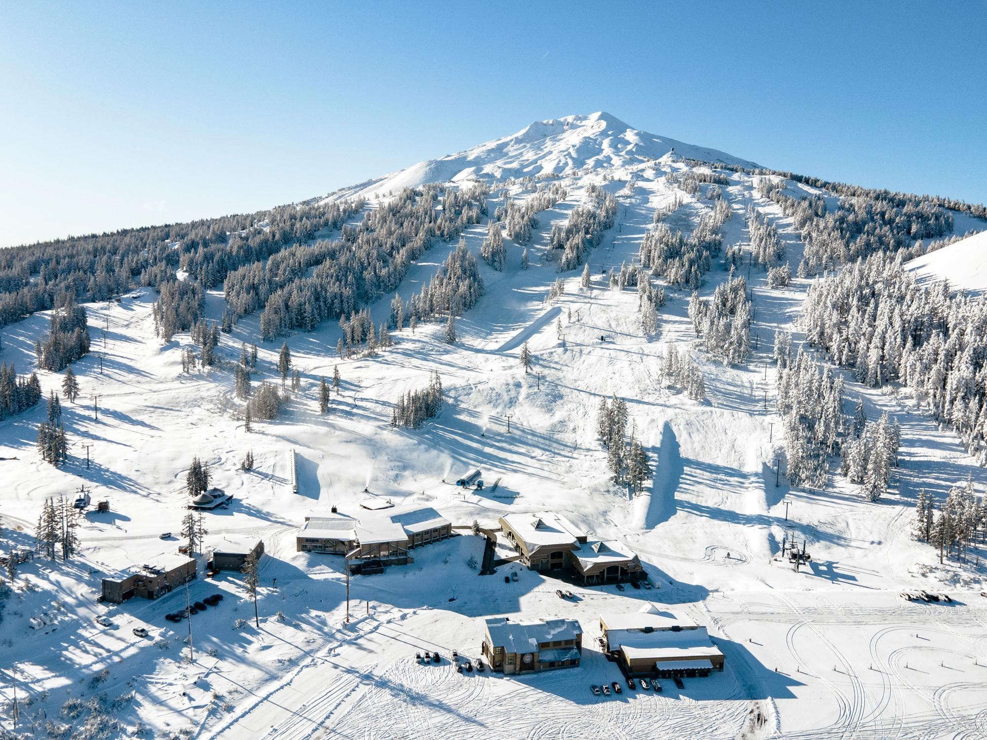 A snowy mountain with ski slopes, wooded areas, and buildings at the base under a clear blue sky. Tracks are visible in the snow around the buildings.