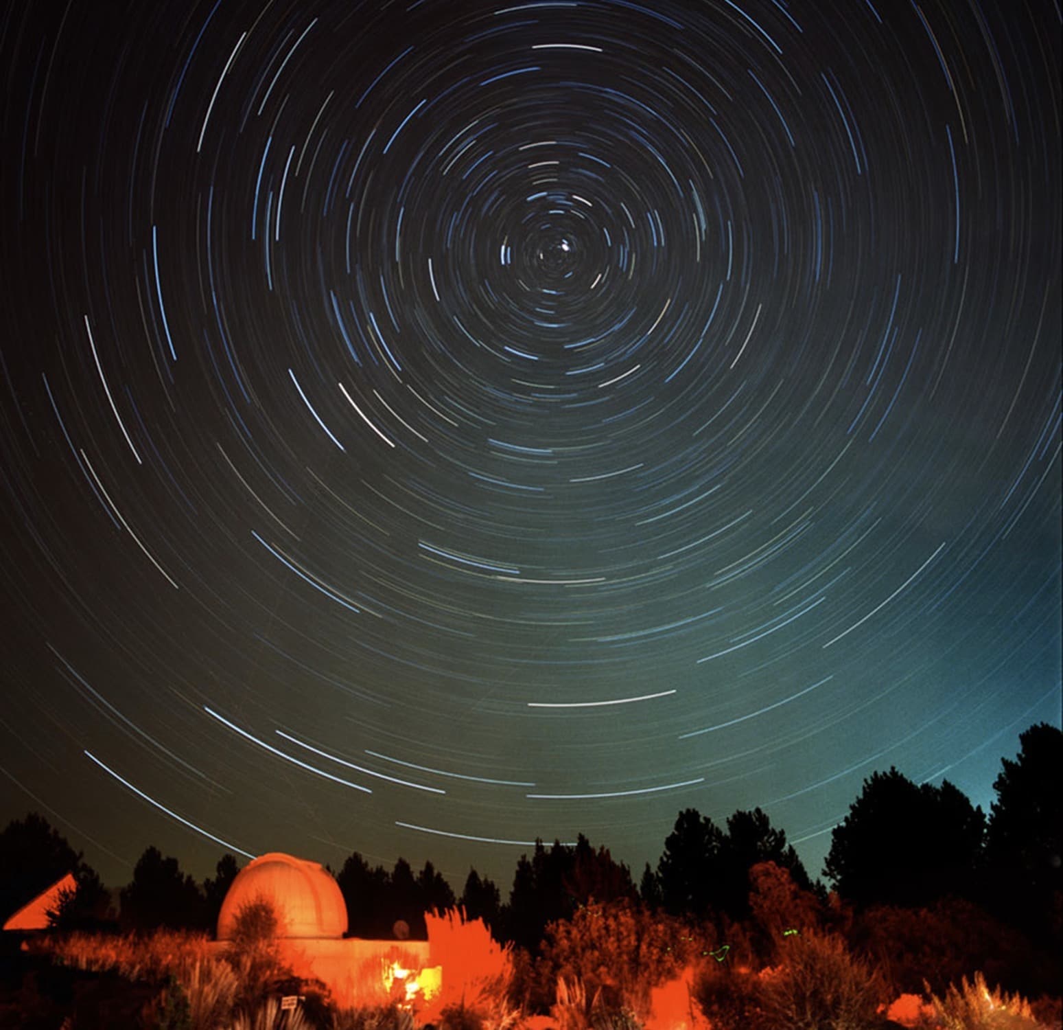 Long-exposure photo of the night sky shows star trails circling above a small observatory dome and trees, with a faint orange glow illuminating the ground.