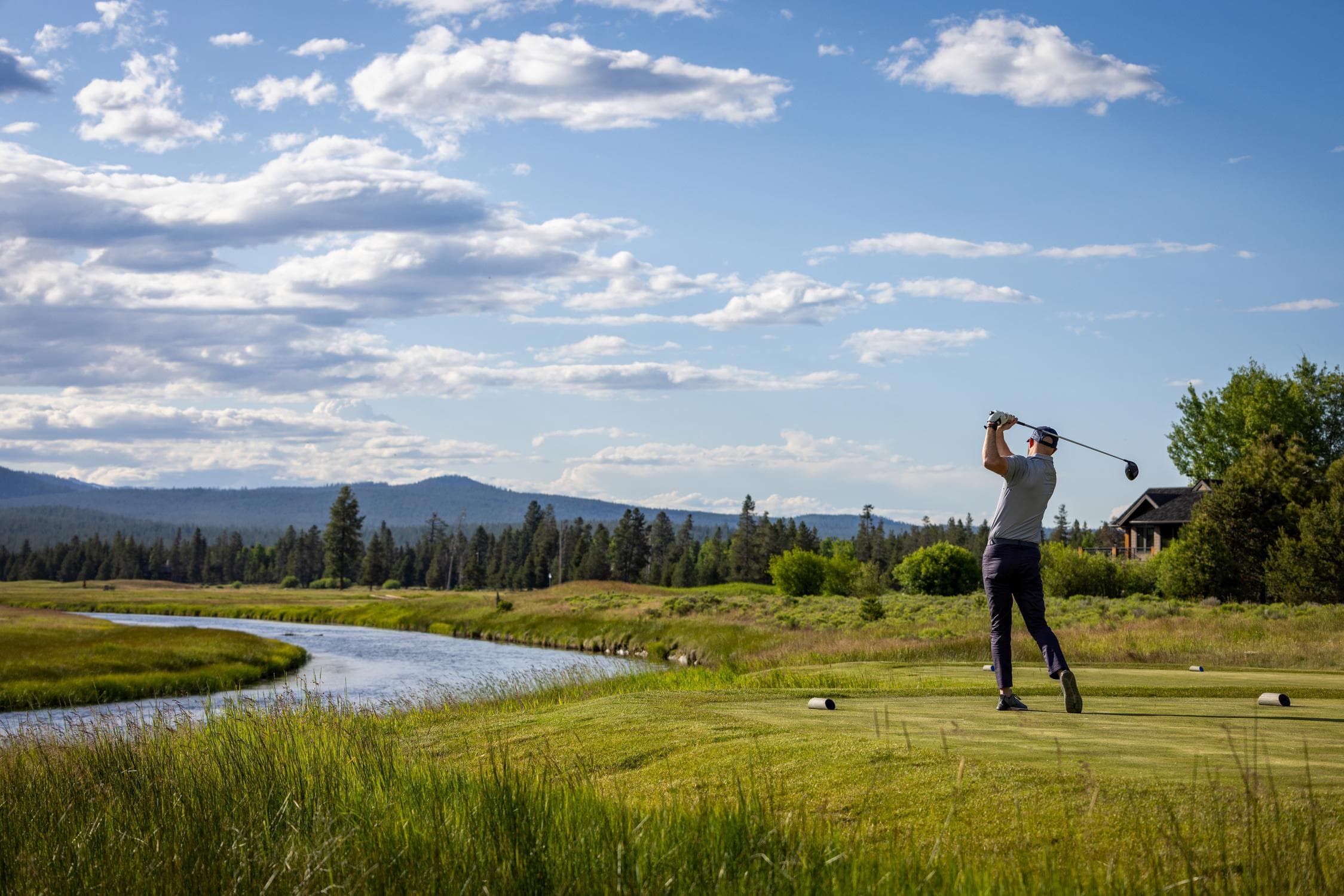 A person swings a golf club on a grassy course next to a winding river, with distant mountains and a partly cloudy sky in the background.