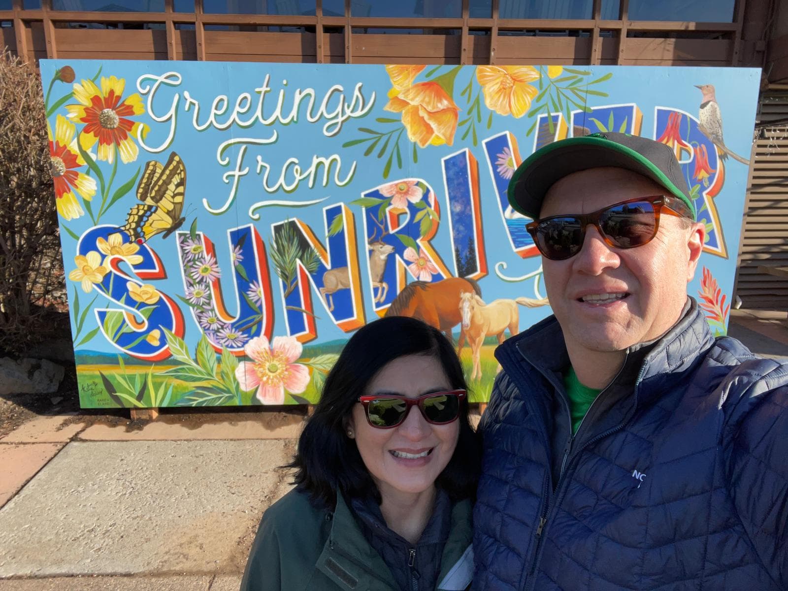 A smiling man and woman in sunglasses pose in front of a colorful sign that reads Greetings from Sunriver, decorated with flowers, a horse, birds, and butterflies.