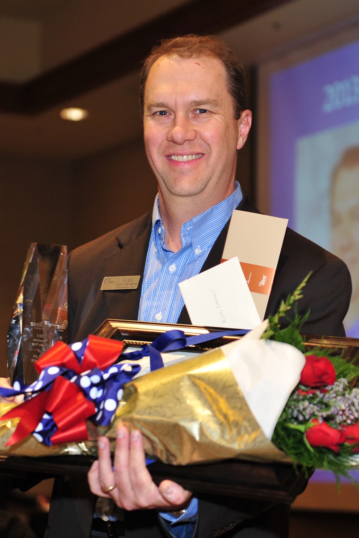 A man in a suit smiles while holding a bouquet of flowers, a glass award, and envelopes, standing indoors at an event.