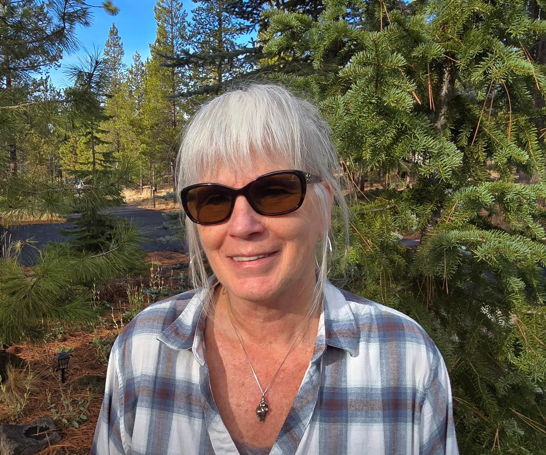 Smiling woman with gray hair and sunglasses stands outdoors among green pine trees, wearing a plaid shirt and a necklace, under a clear blue sky.