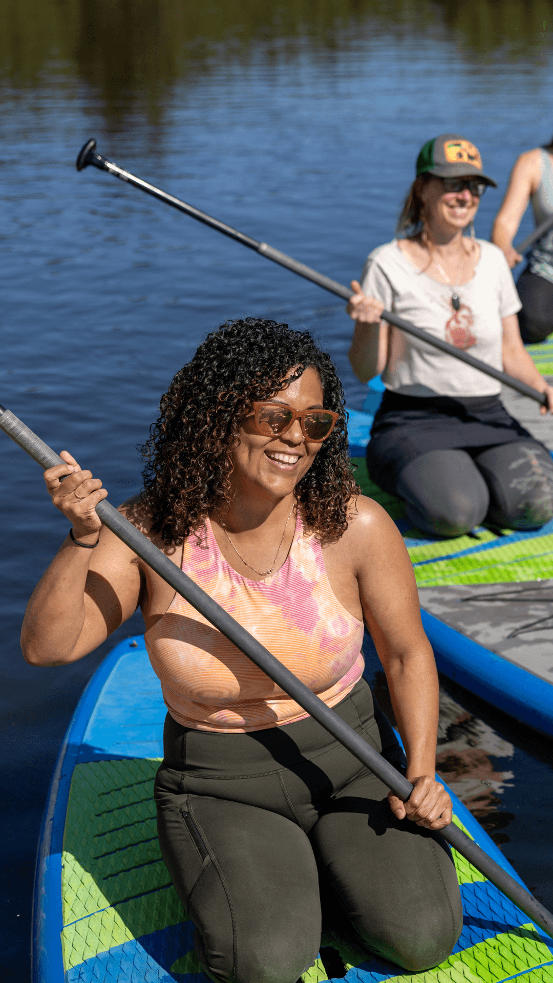 Two women smiling and paddling while sitting on paddleboards on a calm body of water, enjoying a sunny day outdoors.