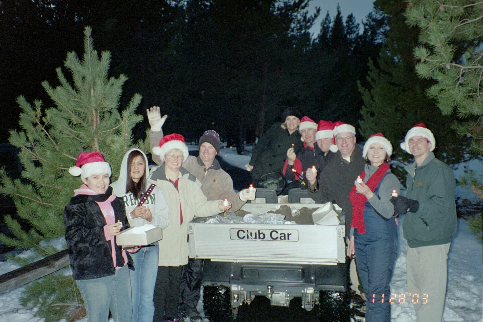 A group of people wearing Santa hats stand around a Club Car outdoors in winter, holding candles and smiling, with snow and pine trees in the background.
