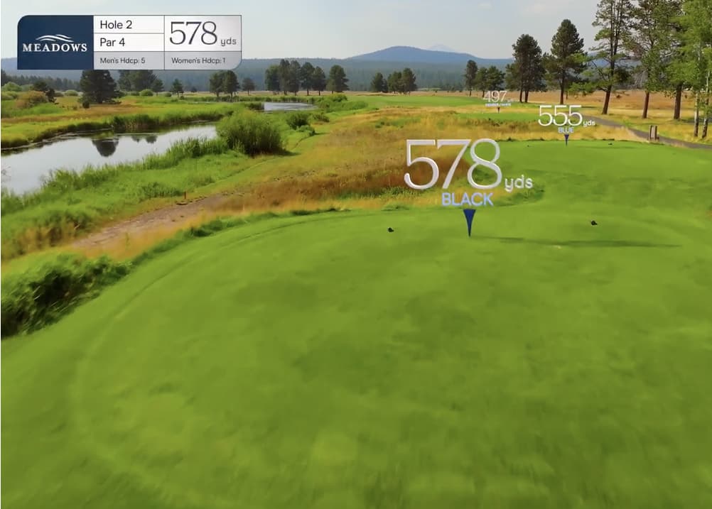 Aerial view of Meadows Golf Course Hole 2, a 578-yard par 4 featuring a water hazard along the left and rolling fairway with distant mountain backdrop.