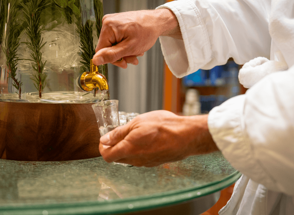 Guest in a spa robe filling a small glass from a self-serve infused water station.