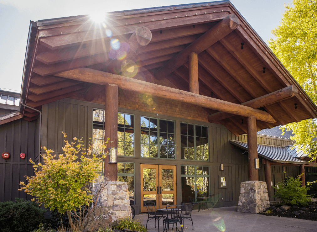 Exterior entrance of a mountain lodge-style spa with timber beams, stone columns, and outdoor seating beneath a sunny sky.