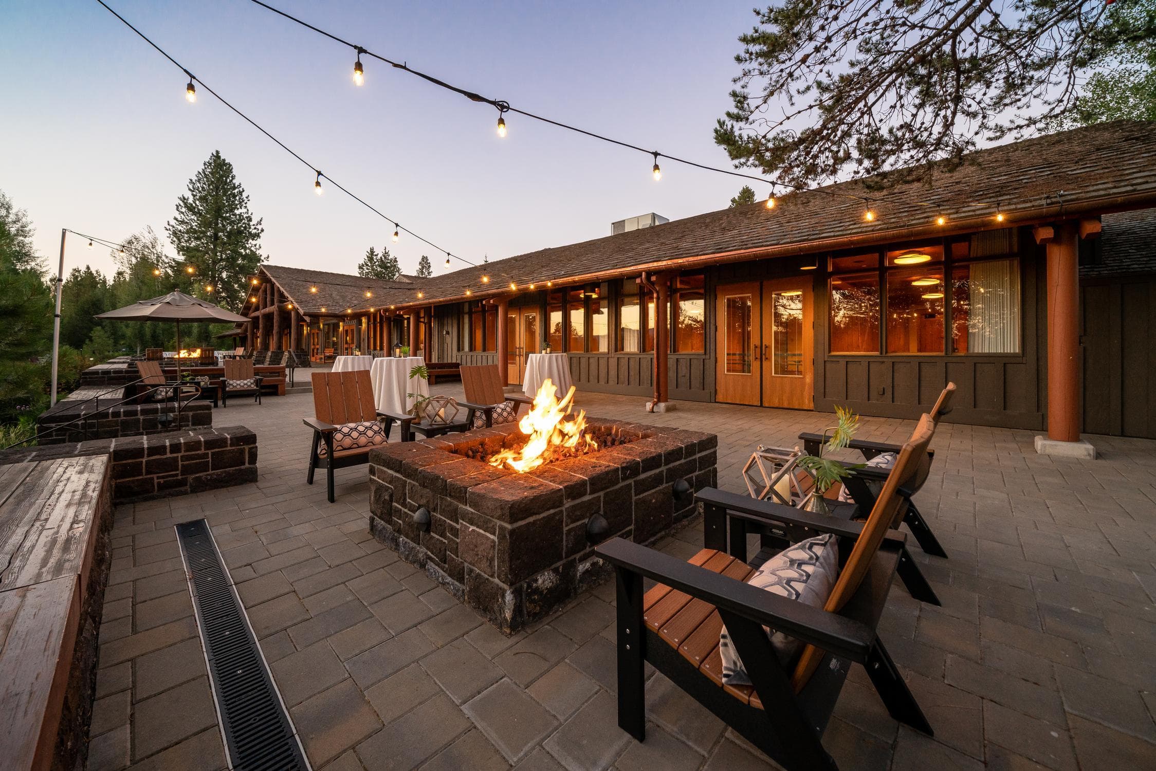 Outdoor patio with string lights, wooden chairs, and a stone fire pit at sunset. The building in the background has large windows and a rustic design, surrounded by trees and outdoor tables.