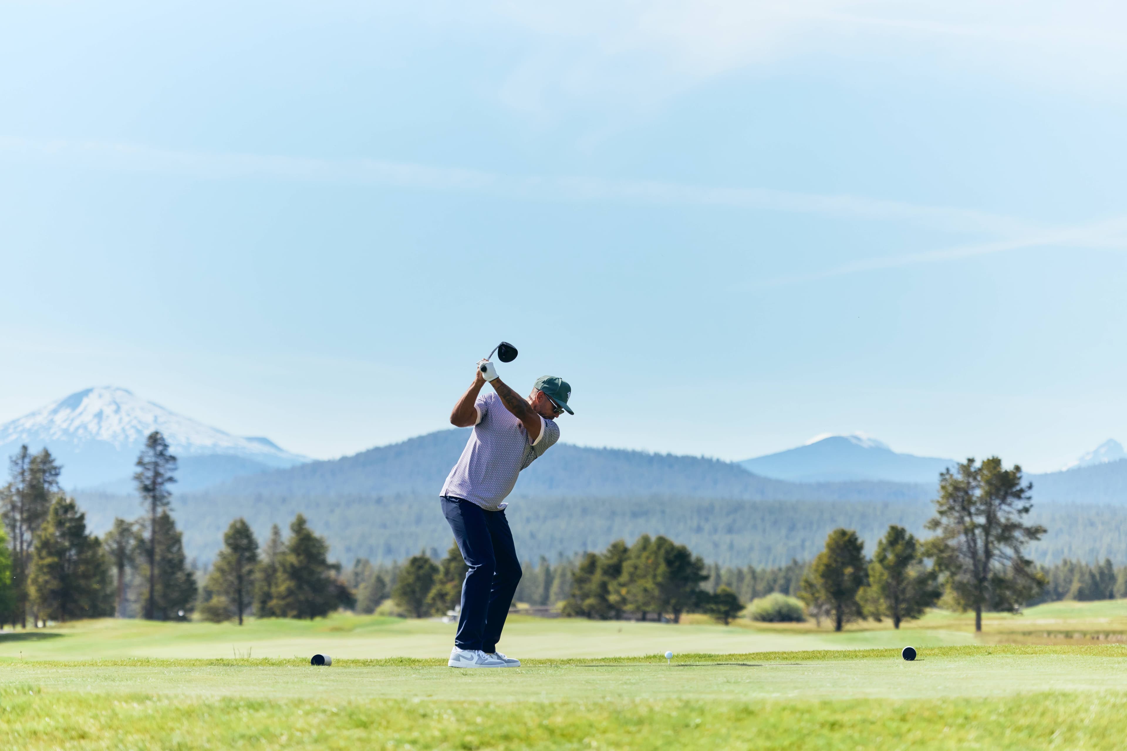 A person is preparing to swing a golf club on Meadows golf course at Sunriver Resort, surrounded by trees and mountains under a clear blue sky.