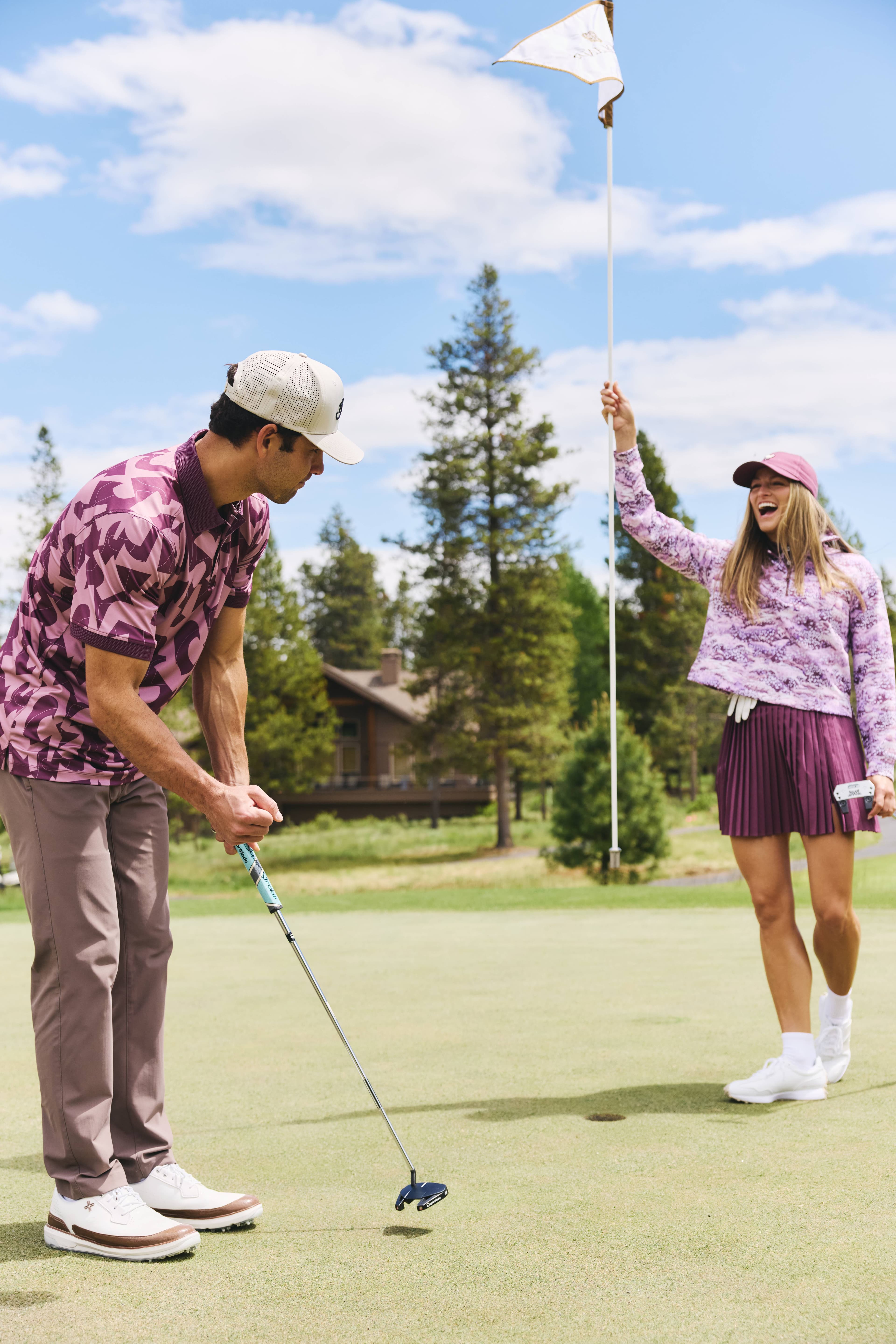 A man in a purple-patterned shirt putts a golf ball while a woman in a purple outfit holds the flagstick and smiles on a sunny golf course with trees and a house in the background on Woodlands golf course at Sunriver Resort.