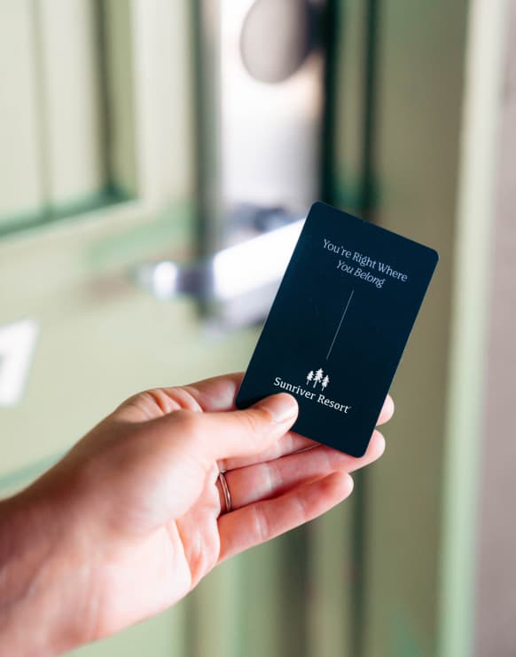 A hand holds a black Sunriver Resort key card in front of a green door. The card reads, Youre right where you belong. The door lock is blurred in the background.