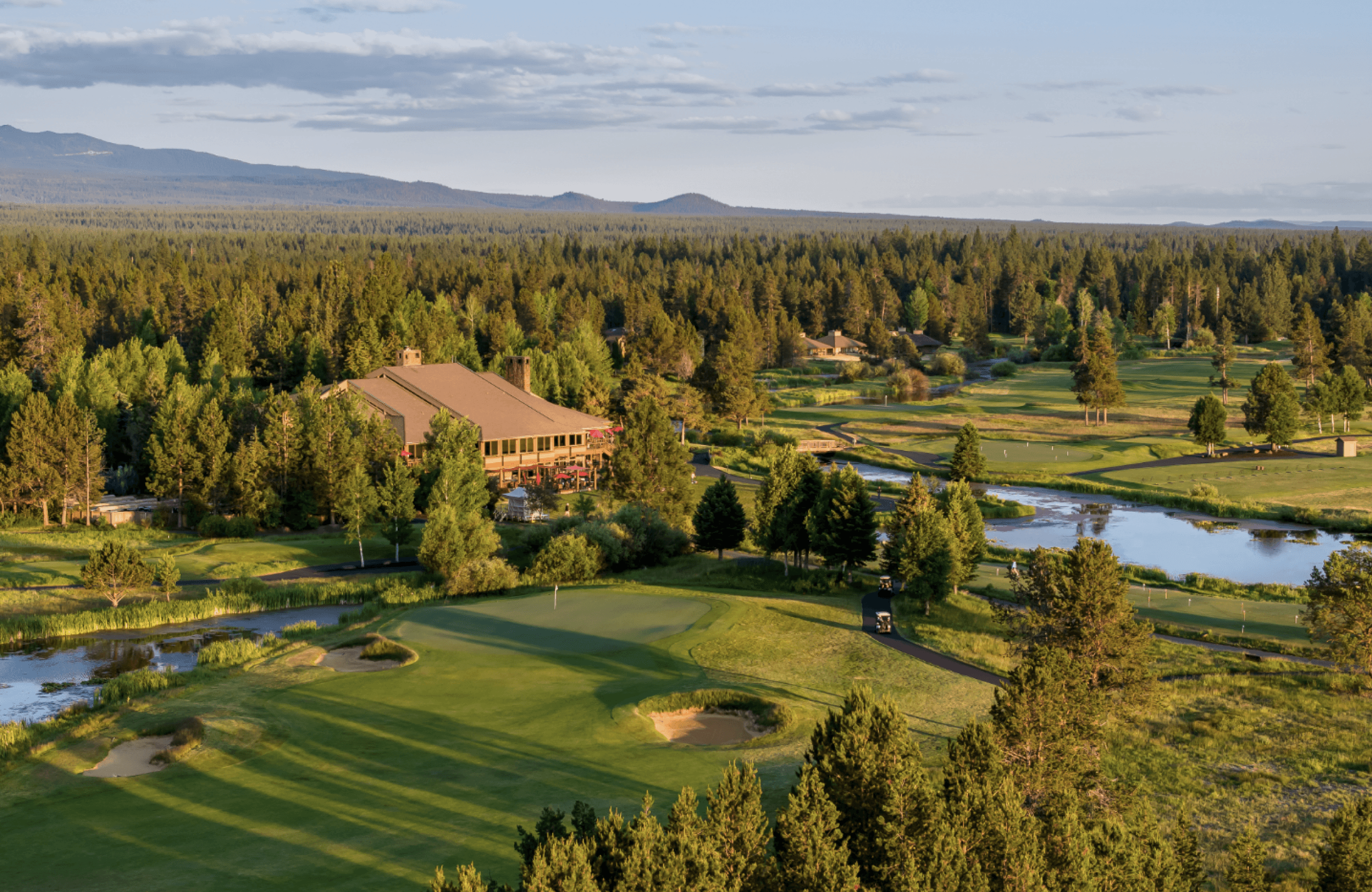 Aerial view of a golf course surrounded by trees, with a large clubhouse in the center, small ponds, sand traps, and distant mountains under a partly cloudy sky.