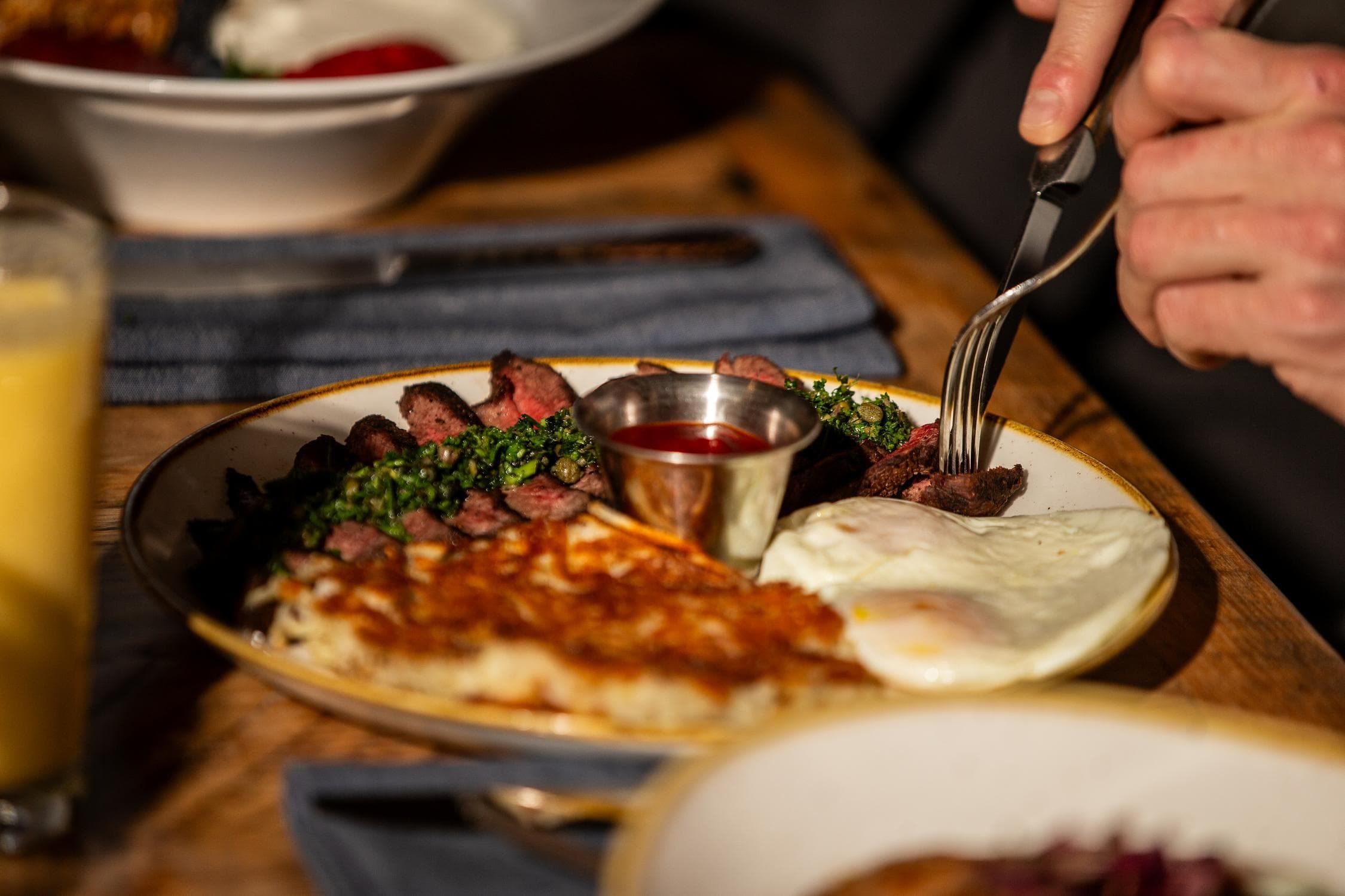 A plate with hash browns, two sunny-side-up eggs, steak pieces, a small cup of sauce, and a green garnish. A person holds a knife and fork, preparing to eat. A glass of orange juice is nearby.
