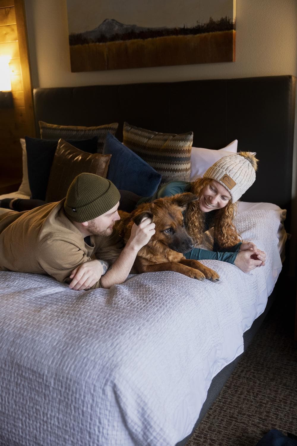 A man and woman in winter hats lie on a bed, smiling and petting a brown dog. Cozy pillows and a painting of a mountain are visible in the background.